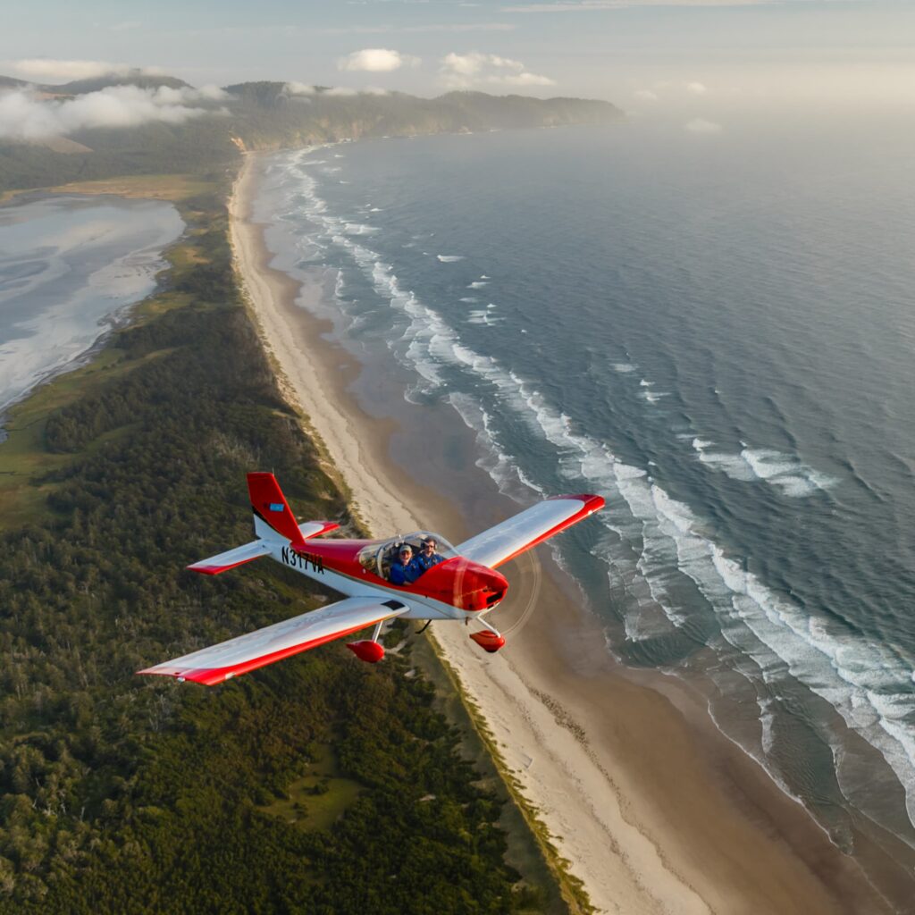 Two people in a red and white plane smile at the camera which flying over a thin stretch of beach.